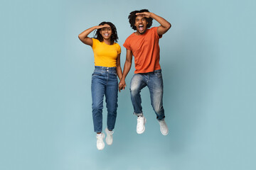 Fototapeta premium Emotional afro-american lovers millennial man and woman in casual posing on blue studio background, bkack couple holding hands, jumping up and looking far away with palm near foreheads, full size shot