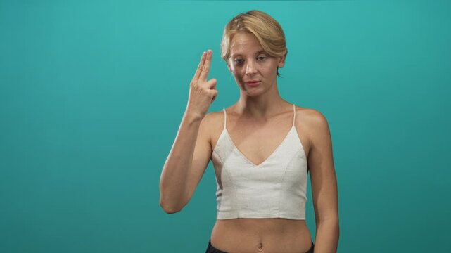 Woman points two fingers to temple, bare navel visible, wearing white crop top against teal studio backdrop; thoughtful contemplation.