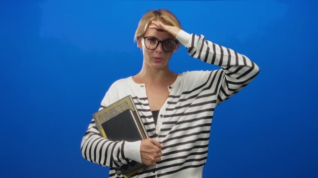 Woman clutching notebooks, hand on forehead shielding eyes, wearing glasses, striped sweater in studio; tired study stress.