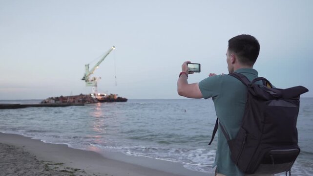 Young traveler blogger takes selfie on phone, makes video broadcast for subscribers on Sea coast about sunken tanker Delf, its rescue using floating crane. Shooting vlog, streaming online podcast.