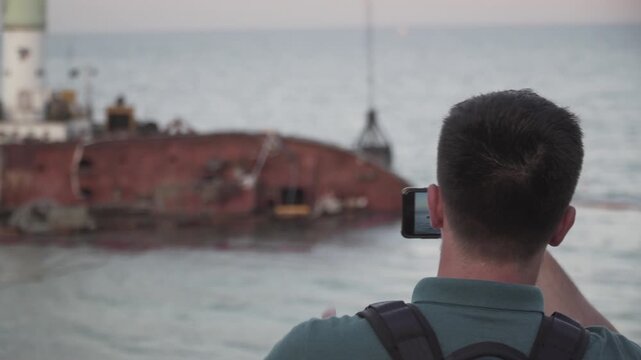 Young traveler blogger takes selfie on phone, makes video broadcast for subscribers on Sea coast about sunken tanker Delf, its rescue using floating crane. Shooting vlog, streaming online podcast.