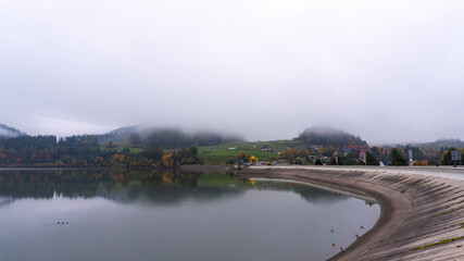 A wide, curved concrete dam stretches across a calm lake, nestled amidst misty rolling hills and a small village under an atmospheric overcast sky. Empty space for text.