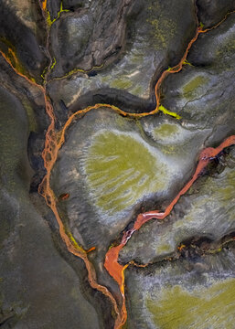 Aerial view of vibrant orange streams meander through a stark landscape of dark rock and patches of bright green moss, creating a stunning natural contrast, Selfoss, Iceland.