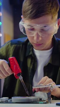 Handsome attantive curious teen boy in protective glasses sitting at the desk soldering on electronic panel at home in the evening. Slow motion.