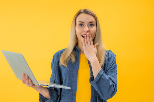 Portrait of a shocked exicted young blonde girl holding laptop computer isolated over yellow background