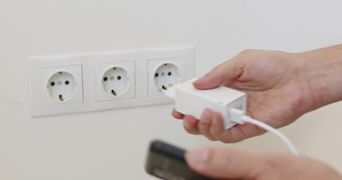 Charger charging smartphone at outlet with cable. Closeup hands connect plug to wall socket to power a device, showing battery top up, electricity use and repair. Neat tech power use idea.