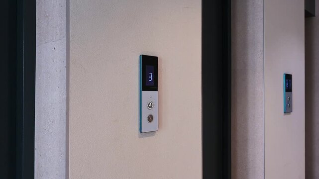 A sleek wall-mounted elevator call panel with a digital display showing floor, two buttons, and a modern blue-gray design; installed in a hallway