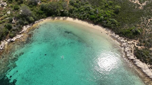 Aerial drone view of a secluded turquoise bay with crystal clear water, sandy beach and rocky coastline surrounded by green Mediterranean vegetation. Summer seascape in Halkidiki, Greece, featuring sh