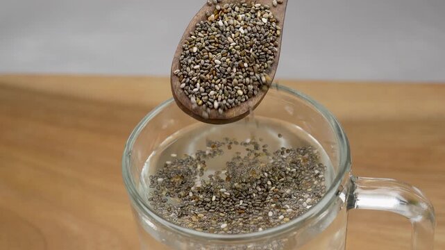Woman adding chia seeds into glass of water on white background. Selective focus on chia seeds.