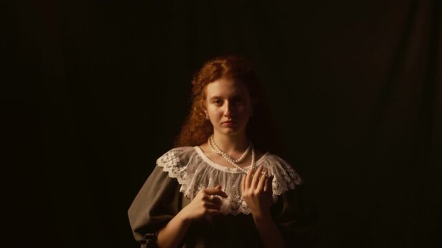 Dolly in shot of red haired young woman in laced dress facing camera while twisting pearl beads of necklace in studio with low key lighting against black backdrop, slow motion