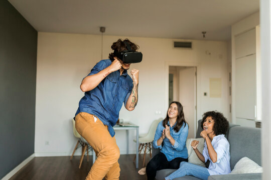Two women encouraging man gaming with VR glasses at home
