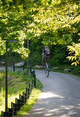 Man riding a unicycle along a sunlit park path surrounded by lush green trees. Eco-friendly urban transport, active lifestyle, balance, sustainability, and outdoor freedom. © Anna