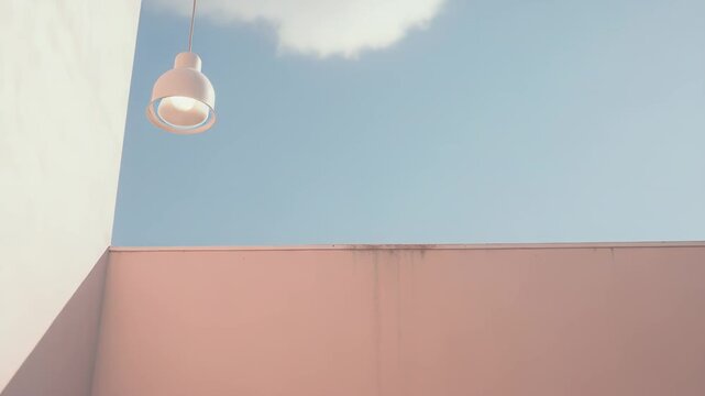 Pale pink minimalist pendant lamp hanging against a soft blue sky with fluffy white clouds and pastel architectural walls, creating a serene and surreal aesthetic