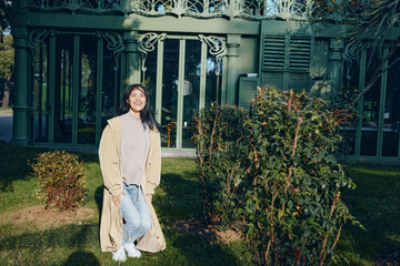 Woman in a long beige coat stands in a garden setting, casual denim and white sneakers, sunny afternoon, greenery and fence backdrop, relaxed lifestyle photo © SHOTPRIME STUDIO