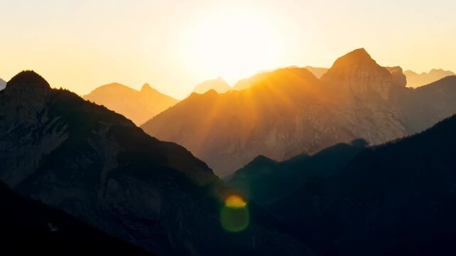 A majestic sunrise illuminates a dramatic, layered mountain range. Golden light rays burst over the peaks, silhouetting the dark slopes and creating a stunning natural spectacle with prominent lens f