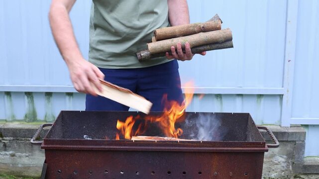 A man adds logs to a roaring fire in a rusty metal grill outdoors, building heat for cooking or warmth.