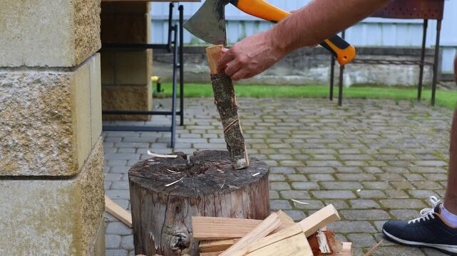 A person splits small logs with an orange-handled axe on a wooden stump in a paved courtyard, preparing firewood for grilling or outdoor cooking.