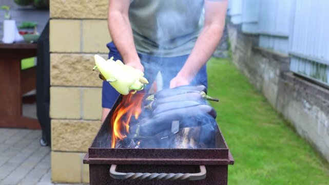 A man grills skewered eggplants and yellow peppers over open flames in a backyard firebox, tending with a long tool amid rising smoke.