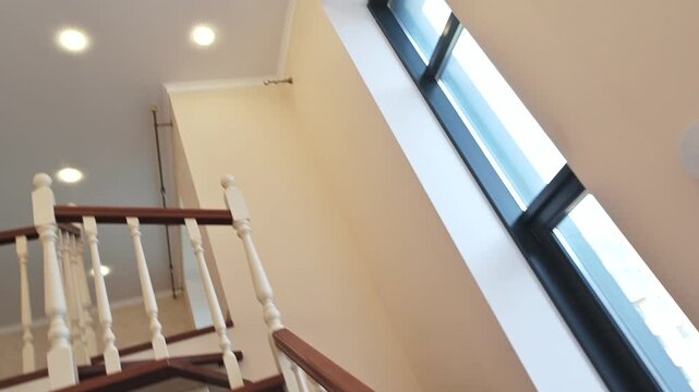 Wooden staircase with white balusters and polished handrail, adjacent to large window; light-colored walls, recessed ceiling light, modern interior feel.
