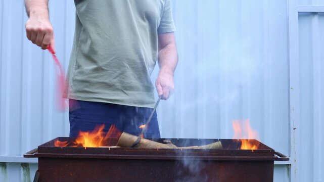 A man adds logs to a roaring fire in a rusty metal grill outdoors, building heat for cooking or warmth.