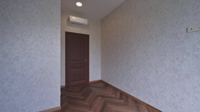 A clean, empty room with light wallpaper, herringbone wooden floor, dark wooden door, ceiling light, and wall-mounted air conditioner &mdash; minimalistic and tidy.
