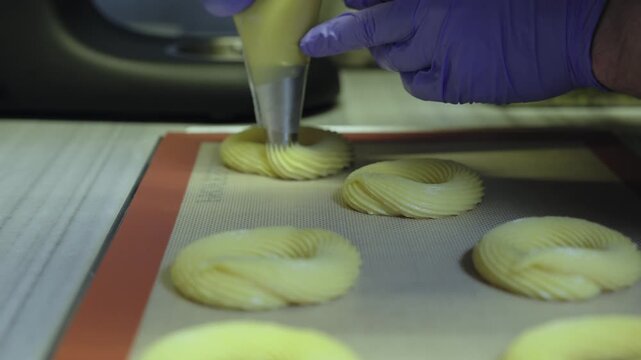 Professional pastry chef piping choux dough for paris-brest dessert onto a silicone mat in a bakery kitchen, preparing traditional french culinary specialty