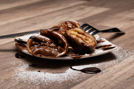 crepe o panqueque relleno con manjar, espolvoreado  con az&uacute;car flor y ba&ntilde;ado en salsa de chocolate, cortado con cuchillo y tenedor