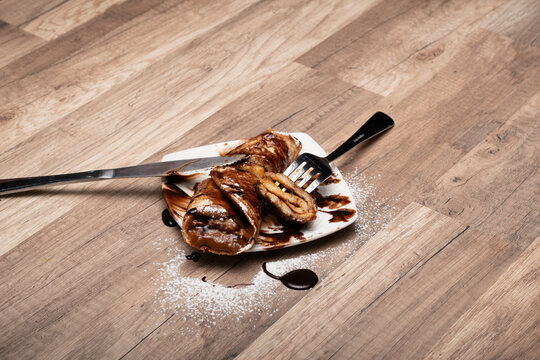 crepe o panqueque relleno con manjar, espolvoreado  con az&uacute;car flor y ba&ntilde;ado en salsa de chocolate, cortado con cuchillo y tenedor