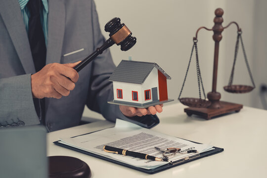 The image depicts a judge's gavel, used for forfeiting property, placed on a table, with a small model of a wooden house behind it, symbolizing real estate law.