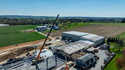 A crane lifts materials at a construction site in a rural location. Workers operate equipment while machines move supplies. Fields and homes are visible in the background under clear blue skies.  © Greg Kelton