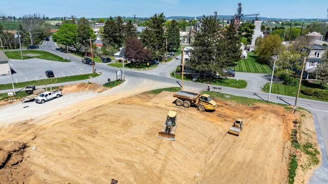 Heavy machinery is present at a construction site in a rural area. Workers use a bulldozer and a grader. Trucks are parked nearby. Trees and buildings are visible in the background.