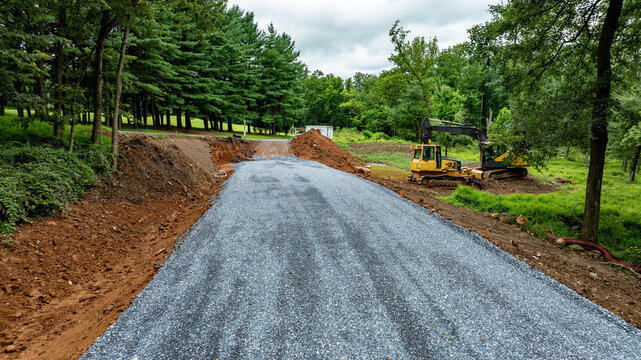 Heavy machinery works on a new road surrounded by trees and dirt piles. The scene shows gravel being laid down under a cloudy sky. Workers are focused on completing the project.