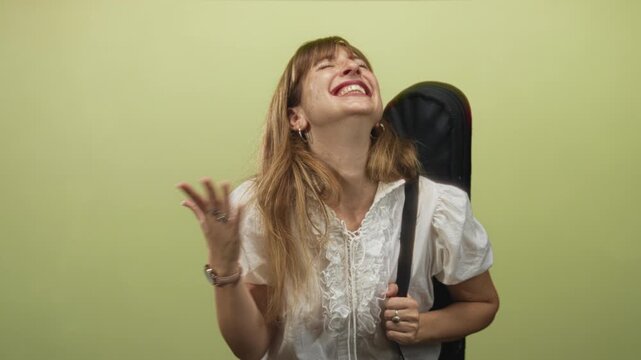 Woman in white blouse holding guitar case on her back with right hand raised and laughing toward camera in studio with lime wall; joy travel adventure.