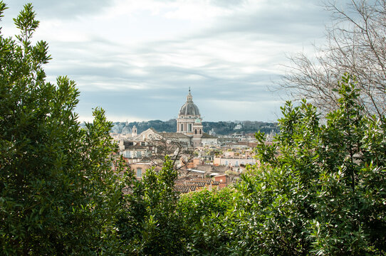 View of Rome skyline with historic church dome framed by lush green trees. Classic European cityscape combining architecture, nature and travel destination appeal.