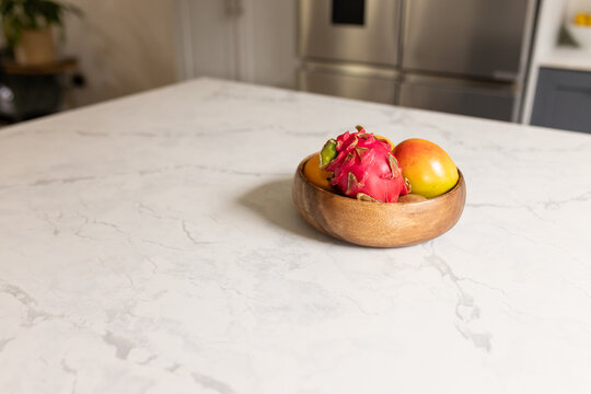Wooden bowl is sitting on marble counter holding dragon fruit and mangoes by fridge, copy space