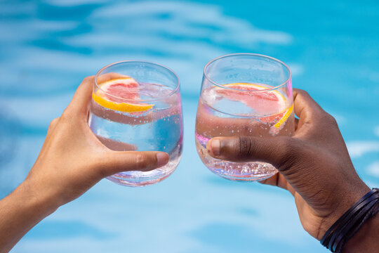 Diverse couple clinking stemless glasses over blue pool with citrus slices bubbles black wristbands