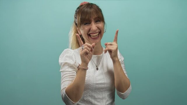Young woman smiling and pointing index fingers upward in studio wearing white eyelet blouse and hoop earrings; joyful playfulness.