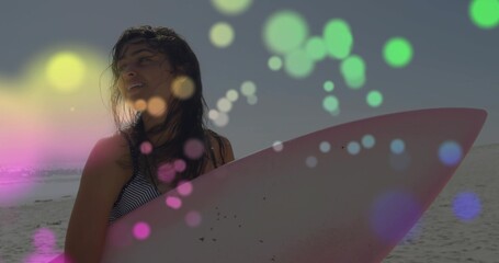 Standing woman holding surfboard on wet sand at dusk, wearing striped swimsuit top, bokeh overlays © vectorfusionart