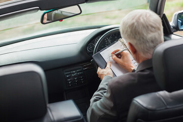 Hands are writing on clipboard in parked car cabin, showing dashboard and grassy roadside