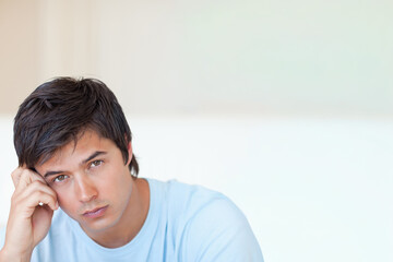 Man in his 20s sitting on studio couch with pale wall wearing light blue T-shirt, gazing