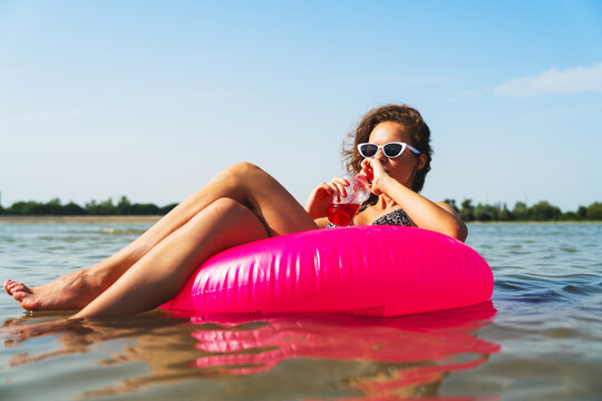 Relaxed woman lounging on floatable swim ring and drinking refreshing drink during summer vacation.