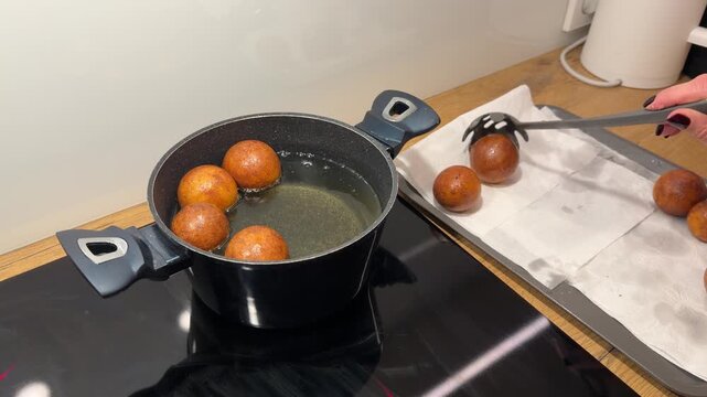 Lifting Fried Dough Balls from Hot Oil to Draining Tray. Hand uses slotted spoon to remove golden-brown dough balls from hot oil. They are placed on paper-lined tray to cool and drain.