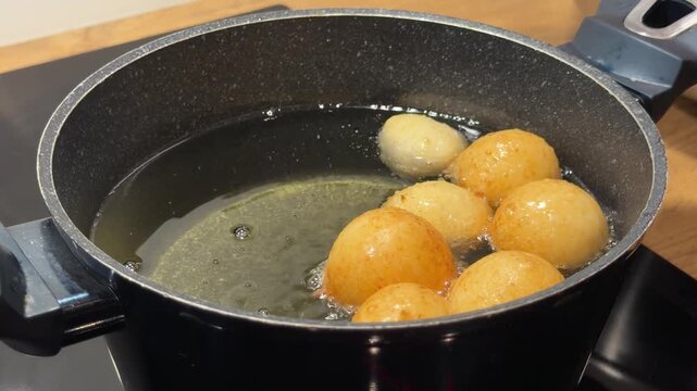 Golden Dough Balls Frying and Bubbling in Oil. Top-down view shows several dough balls at different stages of browning in pot of hot oil. Active bubbling occurs as moisture cooks out of pastry.