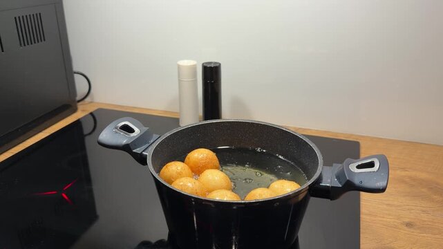 Batch of Dough Balls Sizzling on Induction Cooktop. Black pot filled with golden-brown dough balls sits on modern induction stove. Oil continues to boil around pastries as they finish cooking.