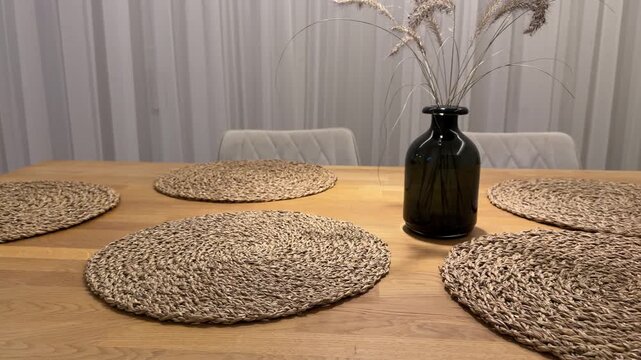 Still Life of Wooden Dining Table with Woven Decor. Detailed view of oak table with circular woven placemats and minimalist vase. Background shows neutral curtain and modern kitchen.