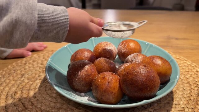 Sifting Powdered Sugar onto Fresh Fried Dough Balls. Child's hand uses small sieve to sprinkle white powdered sugar over plate of golden fried dough balls. Captures final touch of homemade dessert.