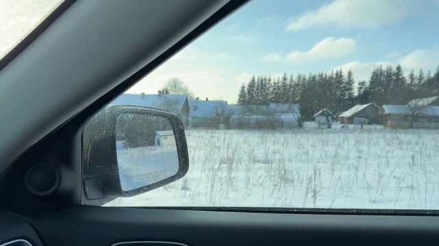Driving Through Snowy Rural Village at Midday. Car travels along snowy road past small houses and open fields under bright winter sky. Roadside lined with tall frosted trees and deep snow drifts.