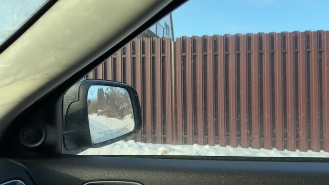 Driving Past Fenced Properties in Winter. View from car window passing residential fences and snow-covered gardens. Modern and traditional houses nestled among frosted trees and blue sky.