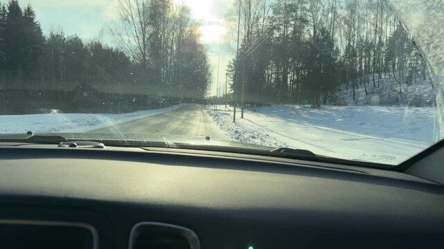 Driving into Sun on Snowy Forest Road. POV from car dashboard driving straight toward bright winter sun on clear road. Tall trees cast long shadows across snow-covered ground on either side.