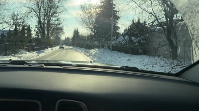POV Driving on Snowy Residential Road with Sun Glare. Driving through quiet residential neighborhood covered in snow. Low winter sun creates bright glare on windshield as vehicle moves past houses.